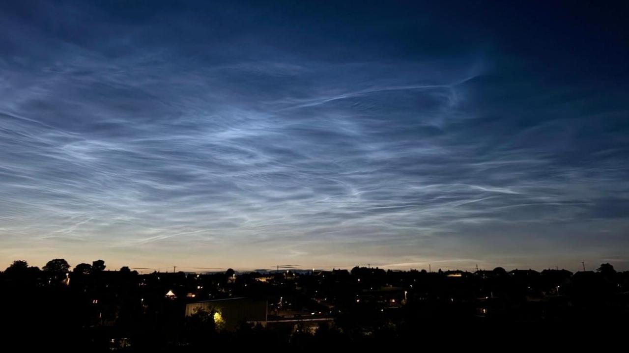 Silver streaks of cloud cut through a navy blue sky. The dark silhouette of a town can be seen below. 