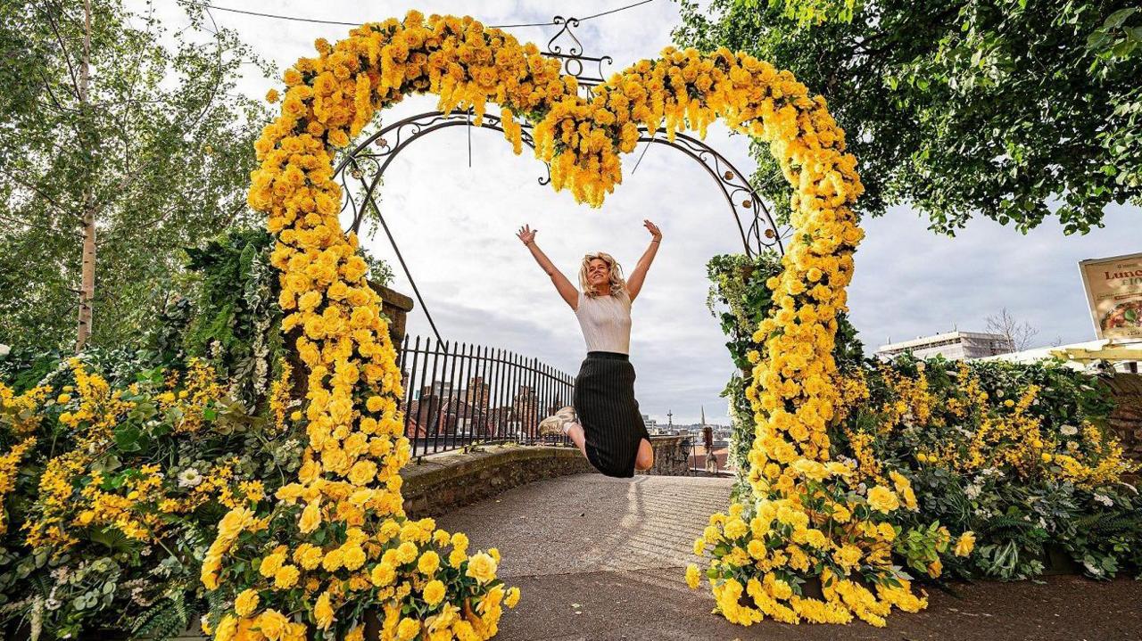 A woman jumps as she is framed by a heart made of yellow flowers at the top of the Christmas Steps in Bristol