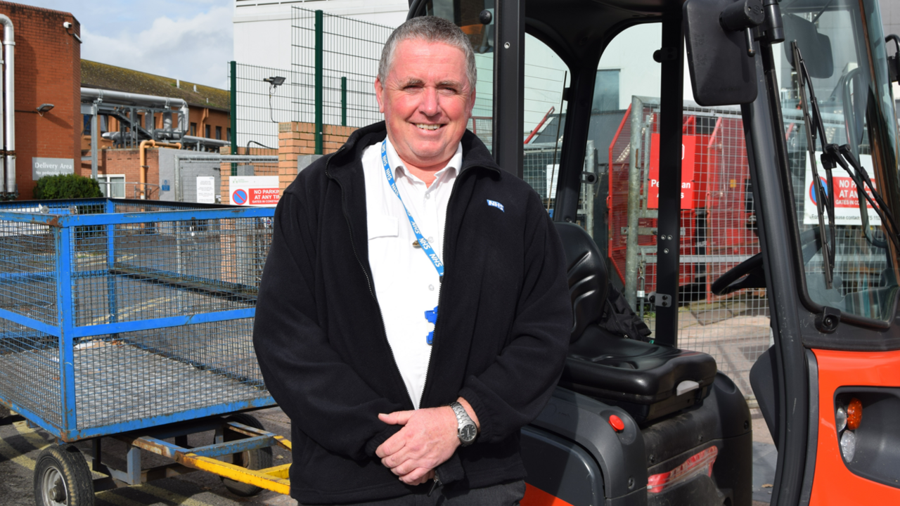 Steve Watts is standing in front of a porter trolley. He is wearing a black fleece and a blue NHS lanyard. He is smiling at the camera.