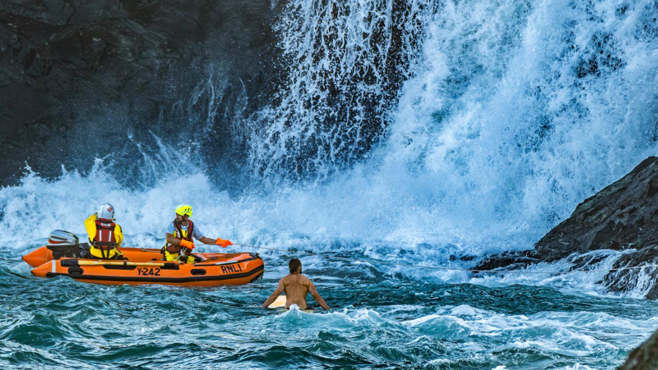 The picture shows a rescue scene with an orange inflatable RNLI rescue boat marked with "Y242" and "RNLI", two rescue personnel aboard the boat, wearing helmets and life jackets and a person in the water, on a surfboard.
Water cascades down rocky cliffs into rough, churning water.