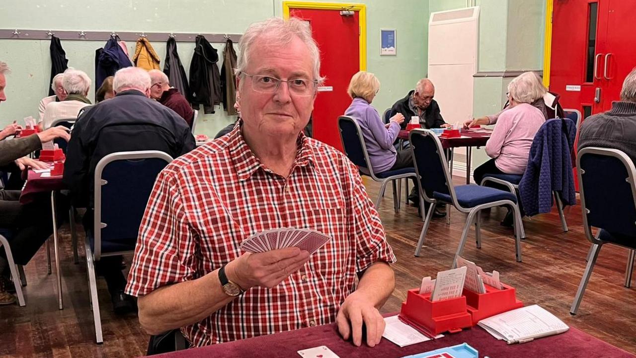 Adrian Darnell playing bridge in a room. He is wearing a red checked shirt, is holding a pack of wards and has items for playing bridge in front of him. He is wearing glasses and has grey short hair. There are other people in the room, behind him, also playing, sitting on chairs at tables. There are coats on hooks, the floor is wooden, the walls are green and there are red doors