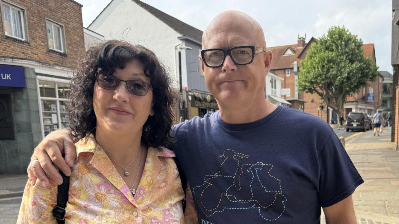 A woman with black, curly hair in a yellow floral shirt and dark glasses standing next to a bald man who has his arm around her and his wearing black glasses and a navy t-shirt. 