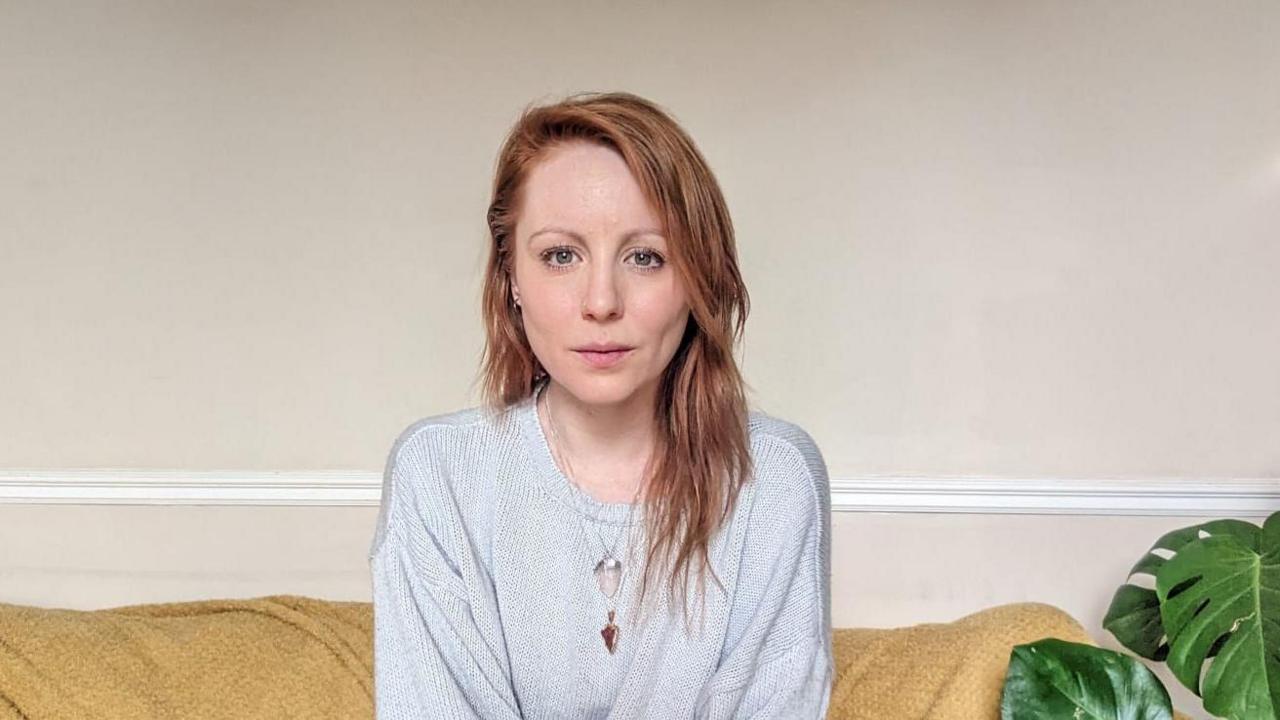 Hannah Clarke, a white woman, sitting on a sofa and wearing a grey top with pink sleeves.