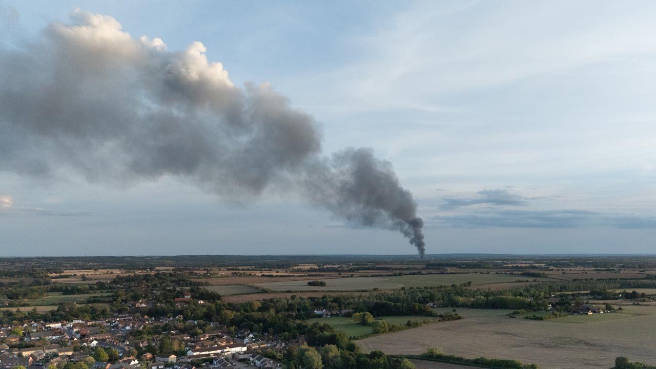 A drone image of black plumes of smoke seen from far away in another area. Houses in a village are in the foreground, before lots of fields which separate the fire from the area.
