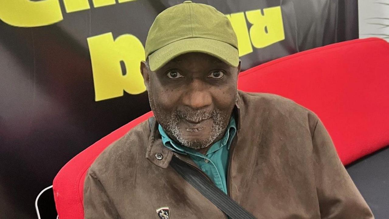 An older man sits on a sofa wearing a green cap and a brown suede jacket. 