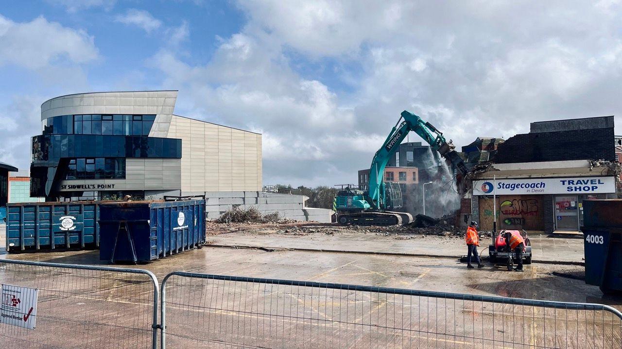 Plant machinery knocking down the old buildings on the former bus station site. Workmen in hi-vis looking at machinery.