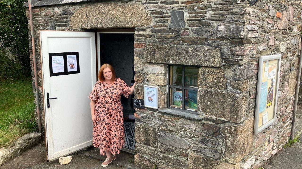 A woman, with a pink dress, standing in the doorway of a small building. The door is open, on the left, and has a poster on it. There is a window on the right and two books on the windowsill.