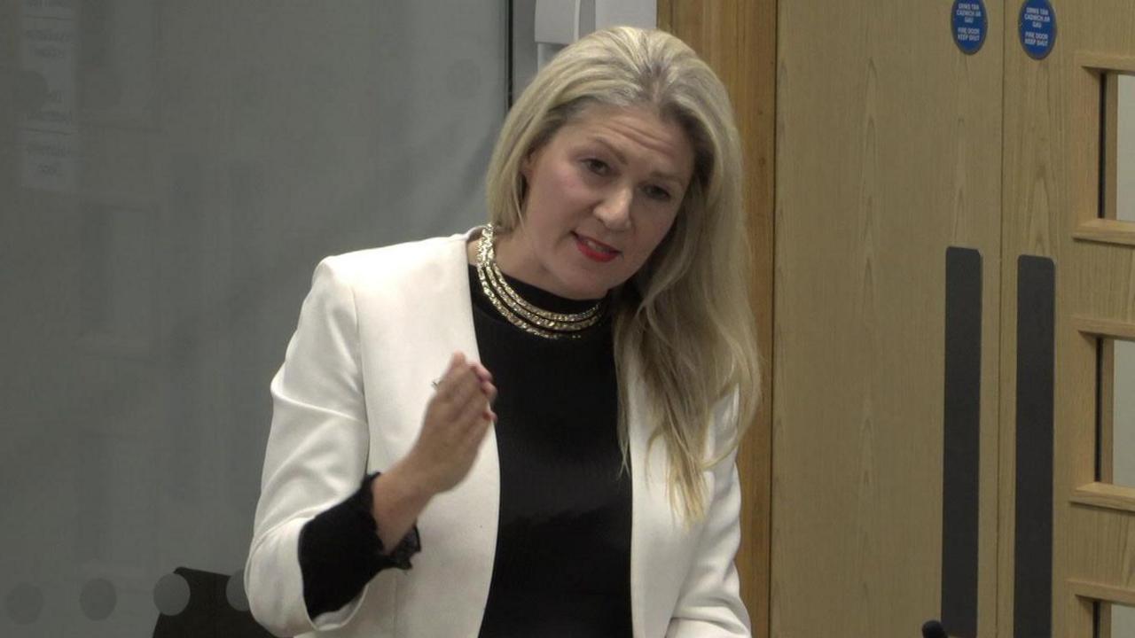 Laura Anne Jones wearing a white jacket and black top, pointing ahead of her with a door visible to the right.