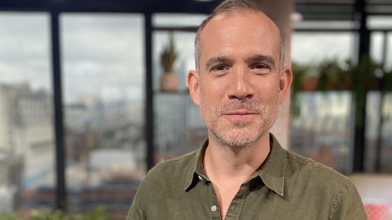 Dr. Xand, a white male with short hair and stubble stands in front of the camera smiling, wearing a dark green shirt