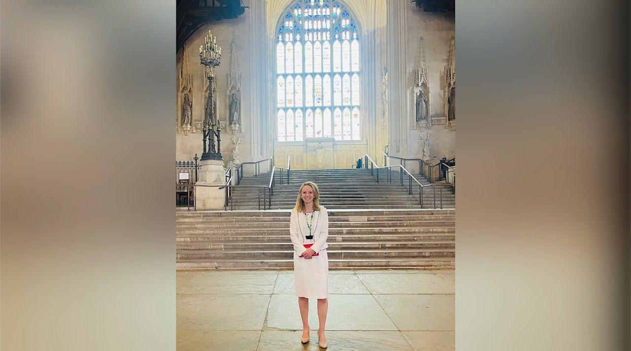 Lucy Rigby with long blonde hair standing in stone-built hall with steps and stained glass window behind her