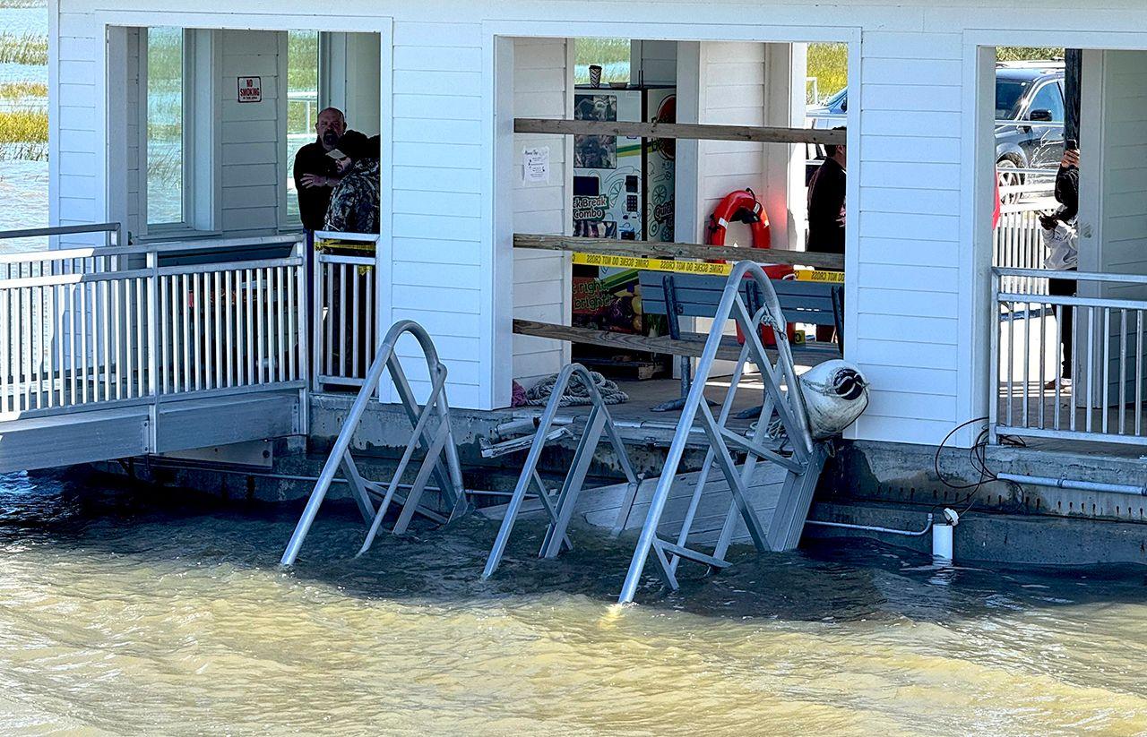 A close up photo shows the collapsed gangway itself, in front of one of the buildings on the dock, with police tape cordoning off the area and a few people looking on.