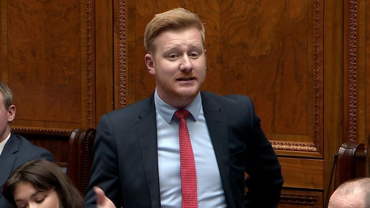 A still of Daniel McCrossan, who has short ginger hair, and is wearing a dark coloured suit jacket, a light blue collared shirt and a red tie. He is mid-speech in the Northern Ireland Assembly, with wood panelled walls behind him.