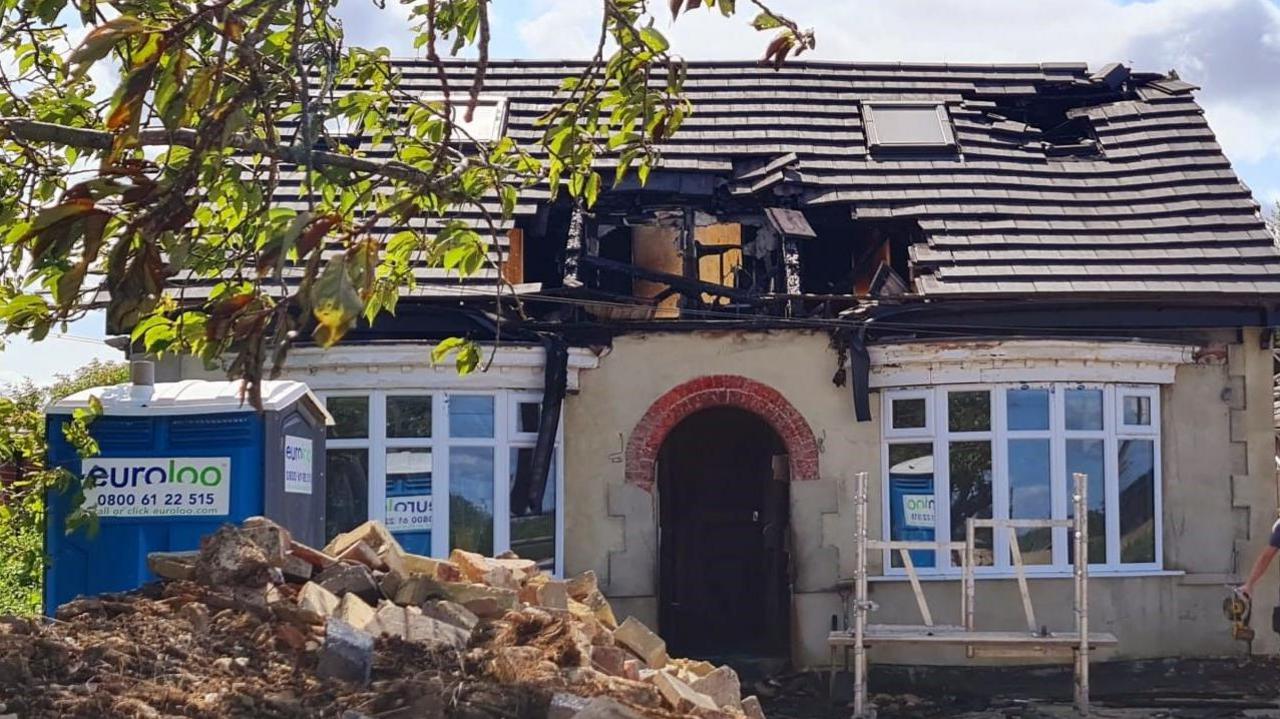 A bungalow with two large bay windows at either side of the property with significant holes in the roof. A pile of rubble sits in front of the property.