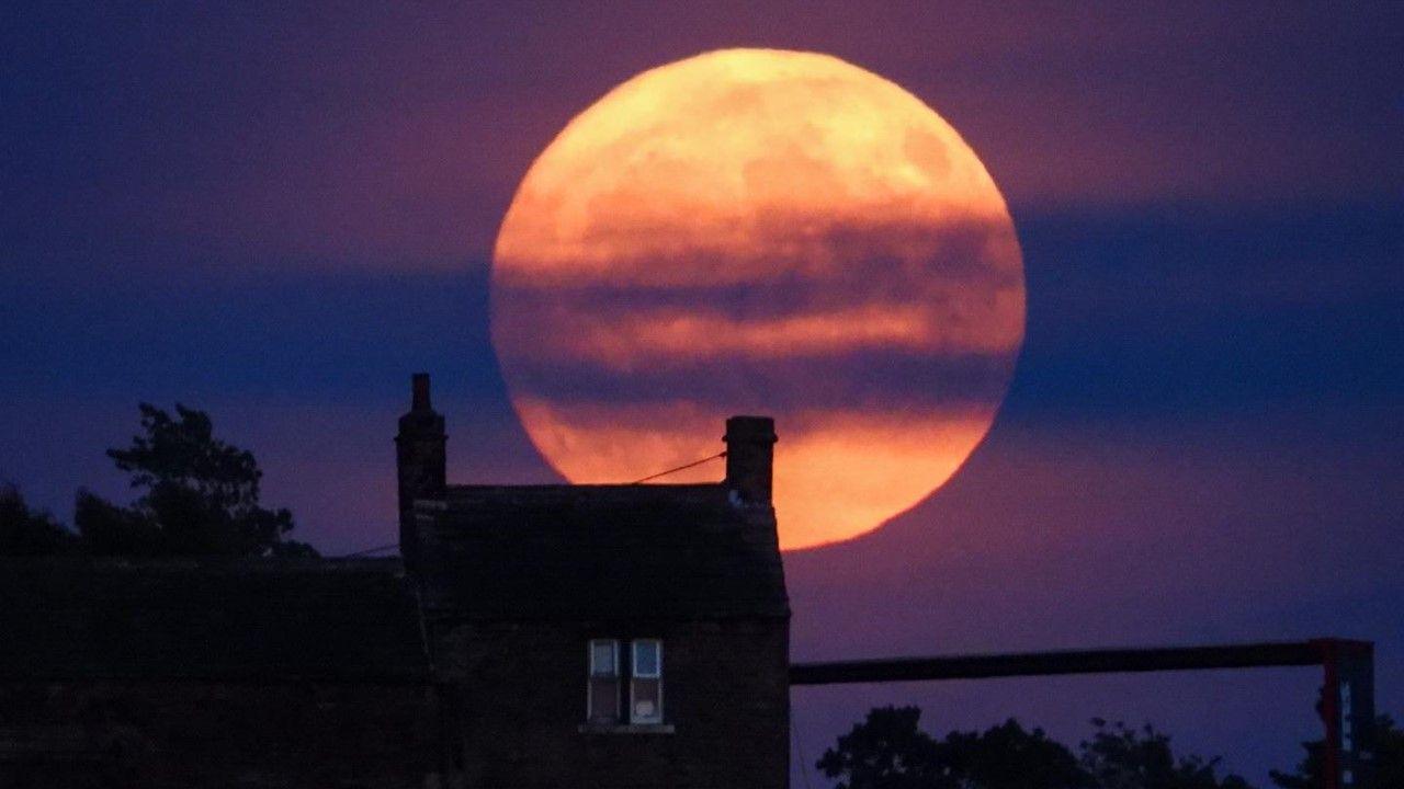 Orange hues of large almost full Strawberry Moon with horizontal purple hue stripes, rises above a house, illuminating a purple and blue sky