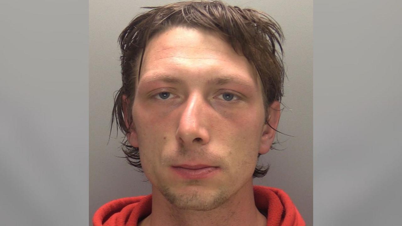 A police mugshot of a man with short dark brown hair wearing an orange sweatshirt. He is looking straight at the camera.