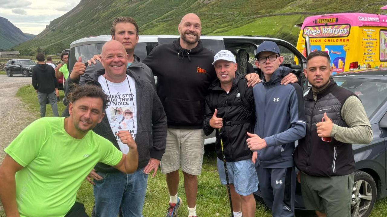 Six men stand in a field and smile with Tyson Fury who stands in the middle of them