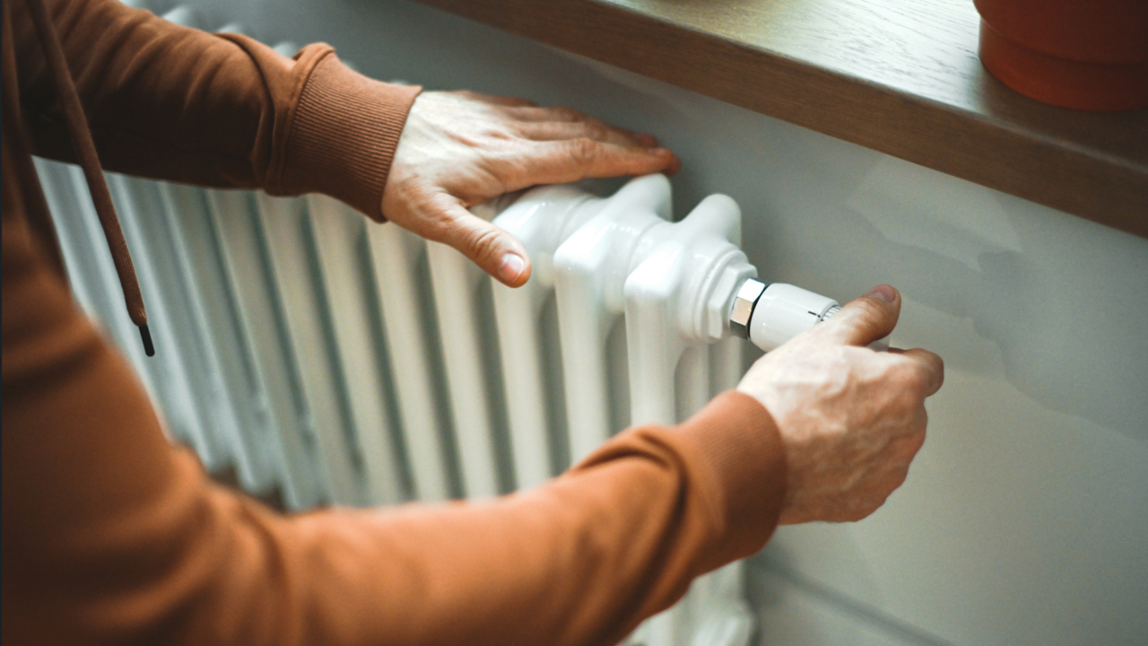 A close-up image of a radiator, there is a person with a brown jumper on touching it. The radiator is white, behind it is a white wall and above it is a brown window sill.