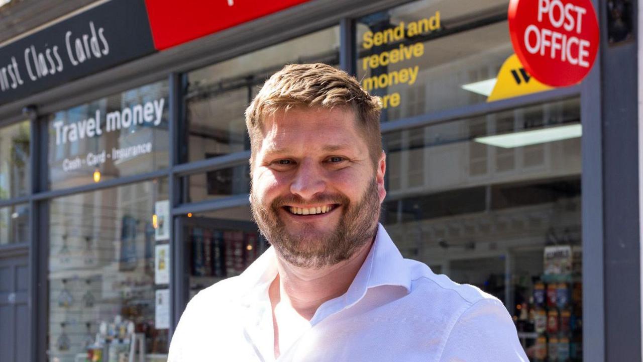 Liam Searle wearing a white shirt and standing in front of a Post Office