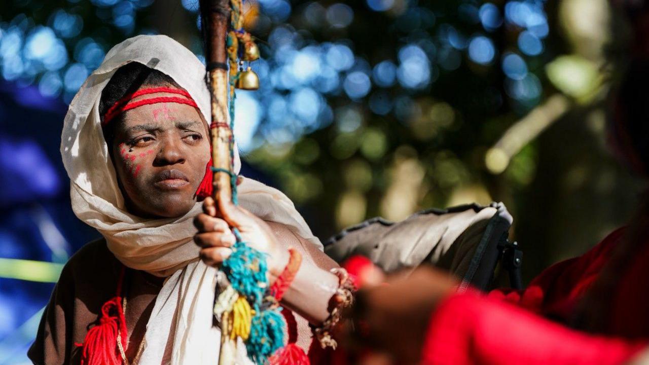 Woman wearing a large white/cream headscarf. She has white and red facepaint and is looking to one side. She is sitting in a woodland setting. 