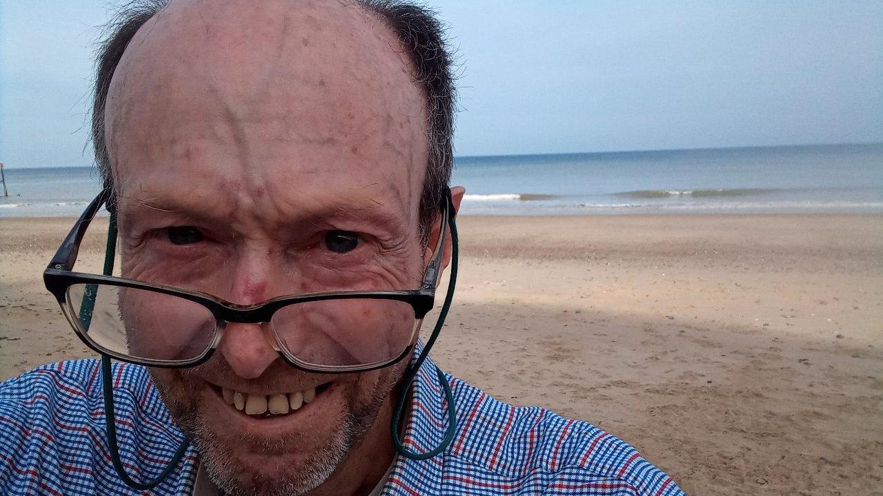 Tim Holt-Wilson takes a selfie style picture. He stands on a beach with the sea behind him. He smiles at the camera and has short dark hair. He wears glasses with a blue shirt. 