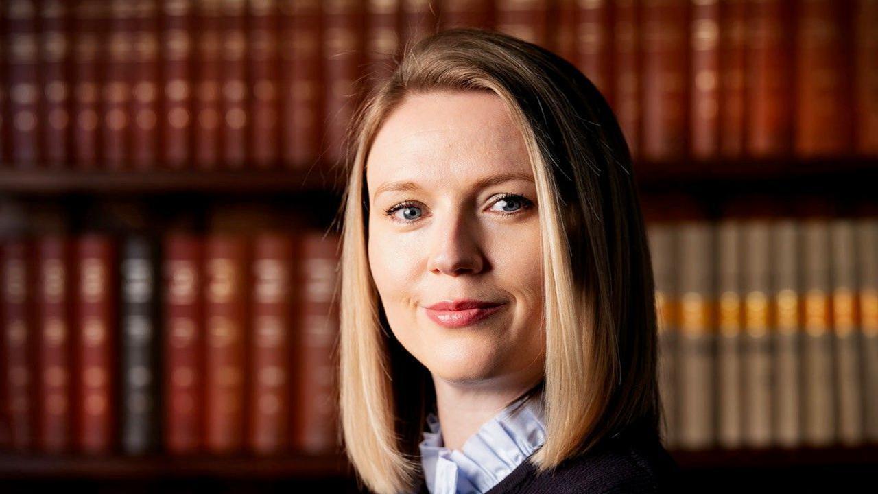 Charlotte Proudman: a woman with a blonde bob hairstyle looking straight ahead. Behind her is a row of red and light-brown books