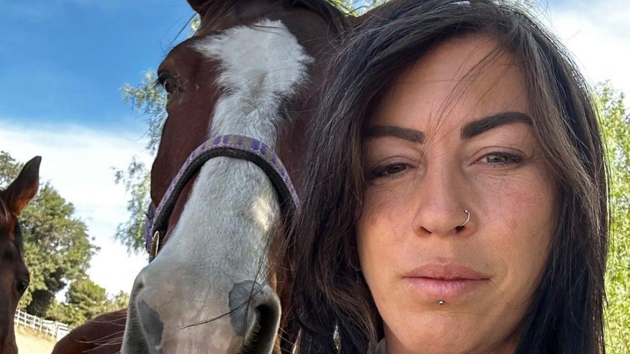 Ms Creasy takes a selfie while standing next to a horse. Ms Creasy has long dark hair and has a nose ring. The horse is a bay colour with a white section stretches from its forehead to its nose.