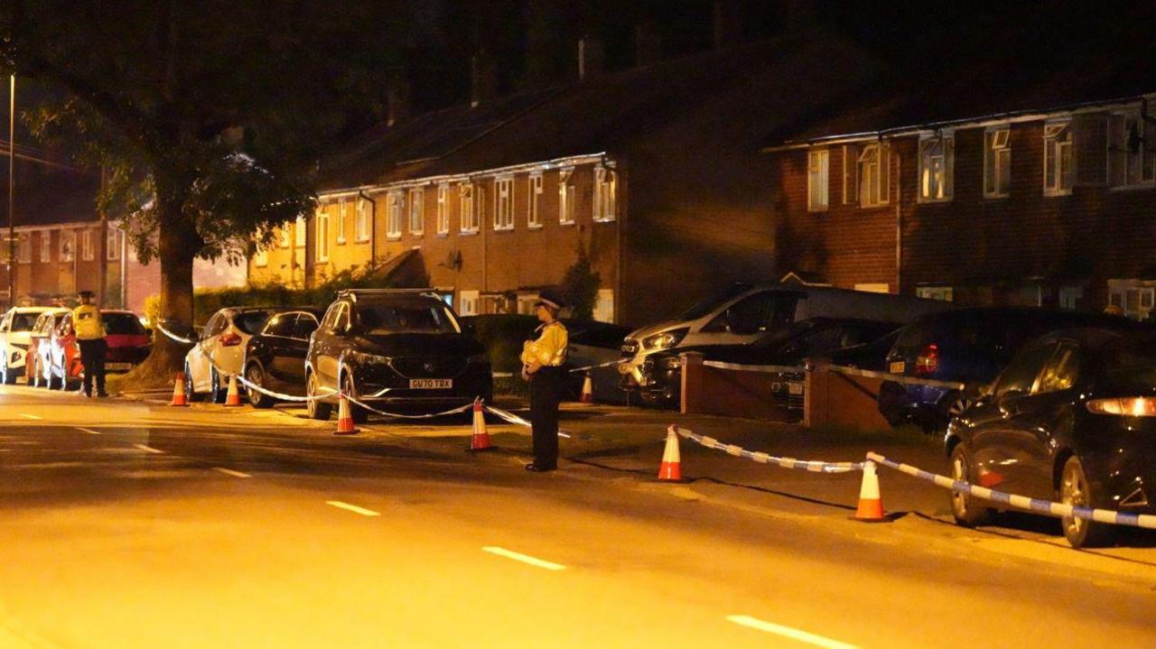 A large amount of police tape cordoning off a number of houses. There are two police officers stood along the cordon, with cars in the background. 