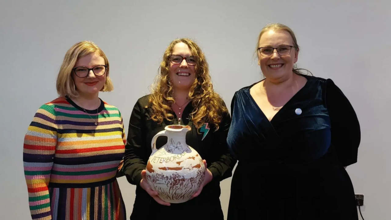 Lizzy Dening, Lauren Kendrick and Hilary Steele standing in a row smiling into the camera. They are standing in front of a white wall. Lauren Kendrick, who is in the middle and has long, red hair, glasses and is wearing a brown top, is holding a large ceramic jug - her award for being named last year's Peterborough Poet Laureate.