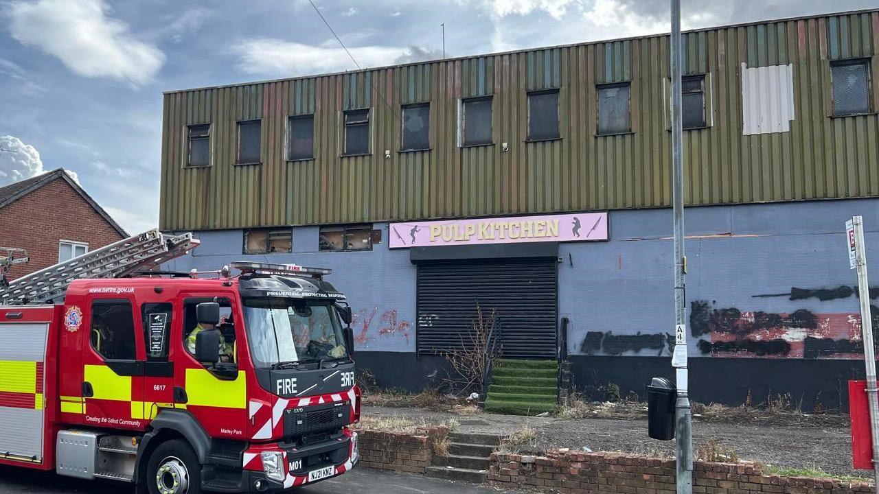 The outside of the Redhouse Working Men's Club - a large building with a corrugated exterior. The small upstairs windows are boarded-up and the main entrance shuttered. Above the entrance is a pink and yellow sign which reads 'Pulp Kitchen'. A fire engine is parked outside.