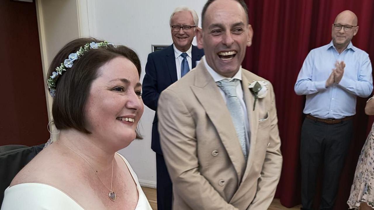 A woman with a white dress and a man with a beige stand in front of a guests while they get married