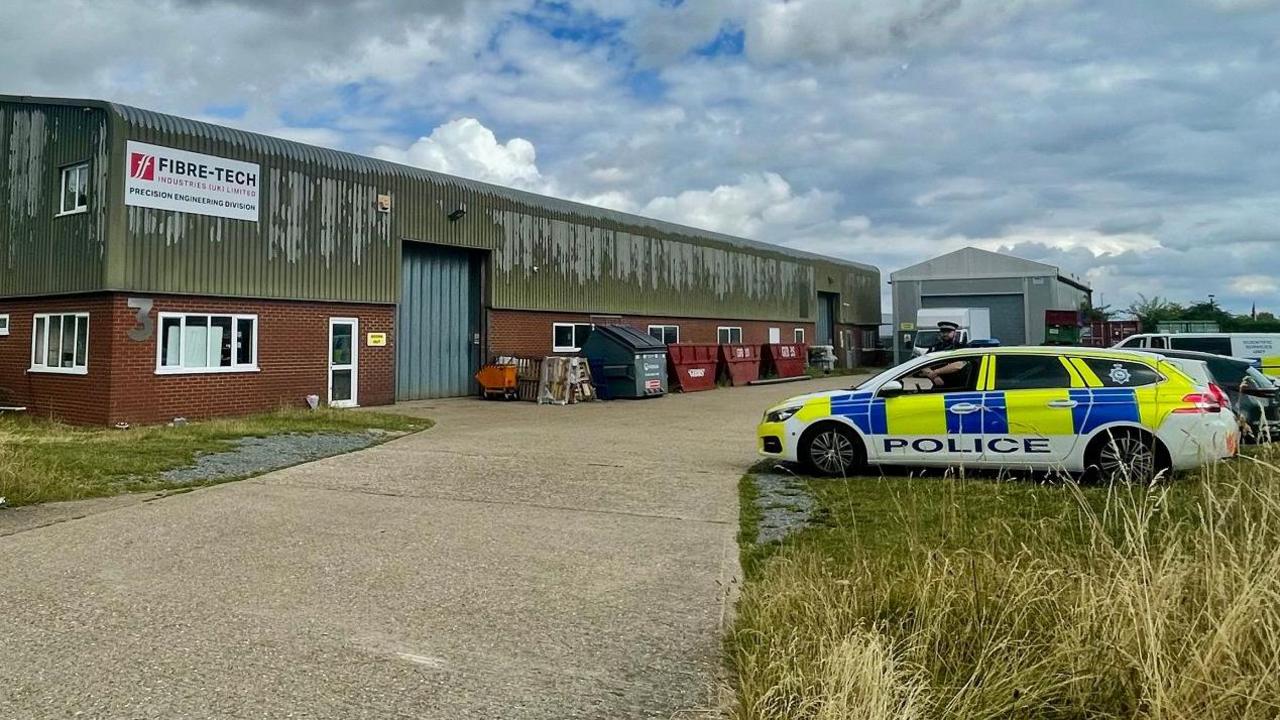 The Fibre-tech warehouse which is brick at the base and corrugated green-grey metal on top. It has its white sign on the side and the two large entrances are closed. A police car is parked on the grass next to the forecourt and an officer stands by it. Another police van is parked further back. A second smaller building like a garage sits next to the warehouse.