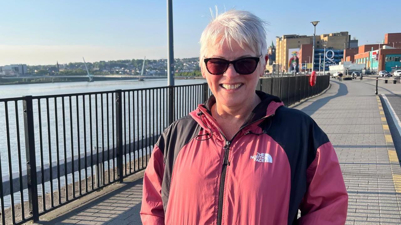 Pauline is smiling at the camera. It is a very sunny day, and she is wearing brown sunglasses. She has short white hair and is wearing a pink 'North Face' coat.