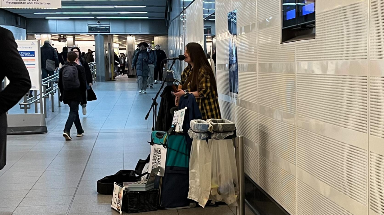 A wide shot of Susana Silva, a woman with long dark hair wearing a black and yellow checked shirt and holding a guitar, singing into a microphone in a passageway at Liverpool Street Station as people pass by her