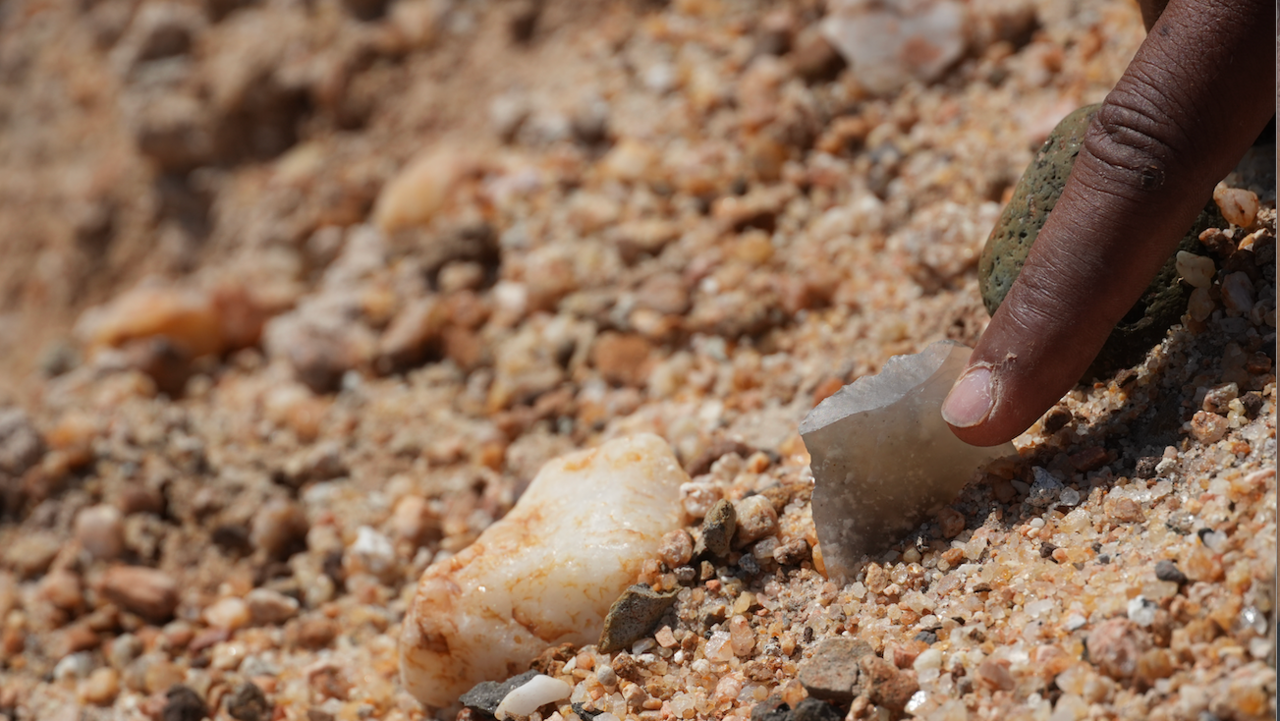 A person's hand is pointing to or holding a sharp stone tool that is partially buried in sandy, gravelly ground. The scene is very close up, showing the texture of the dirt and small rocks around the tool. The focus suggests the discovery or examination of a potential artifact, likely during an archaeological dig.