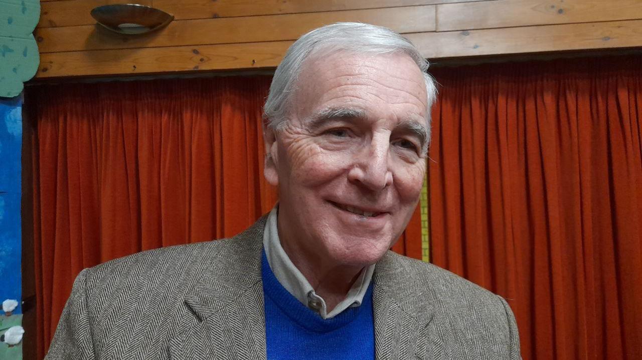 A head-and-shoulders image of Tim Willbond wearing a blue sweatshirt and a grey blazer. He is standing in a hall in front of a red curtain. He is looking at the camera and smiling.