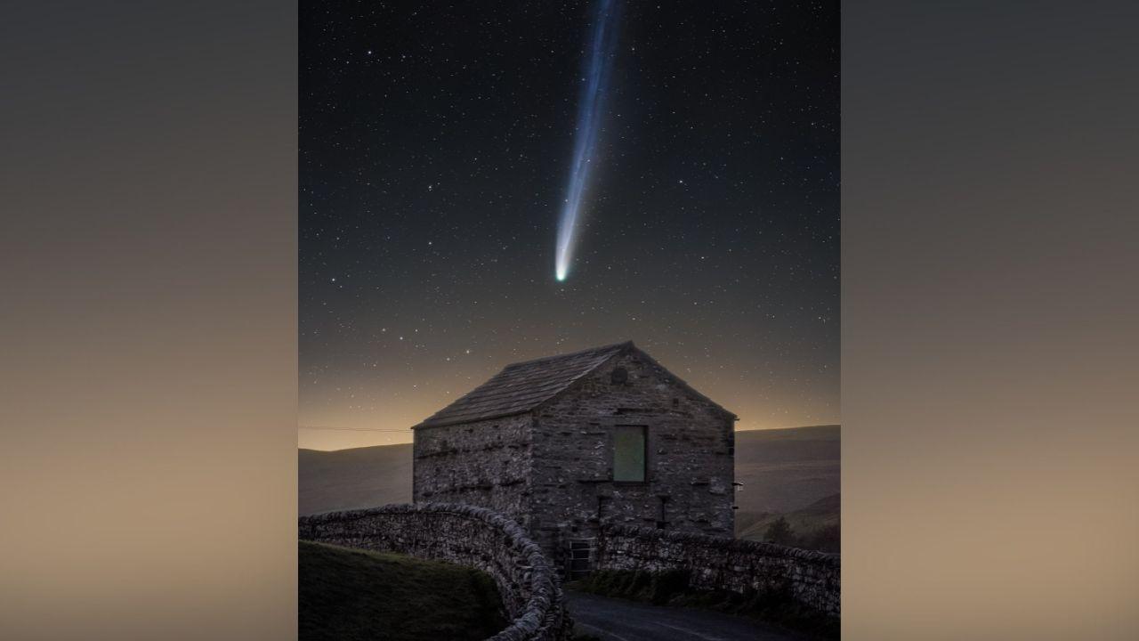 A comet seen in the night sky above a stone house in the Yorkshire Dales. The house is small with just one single window, and is at the side of a road lined by dry stone walls. The comet is a ball of white light in the centre of the photo with a white tail fading to blue towards the top of the picture.