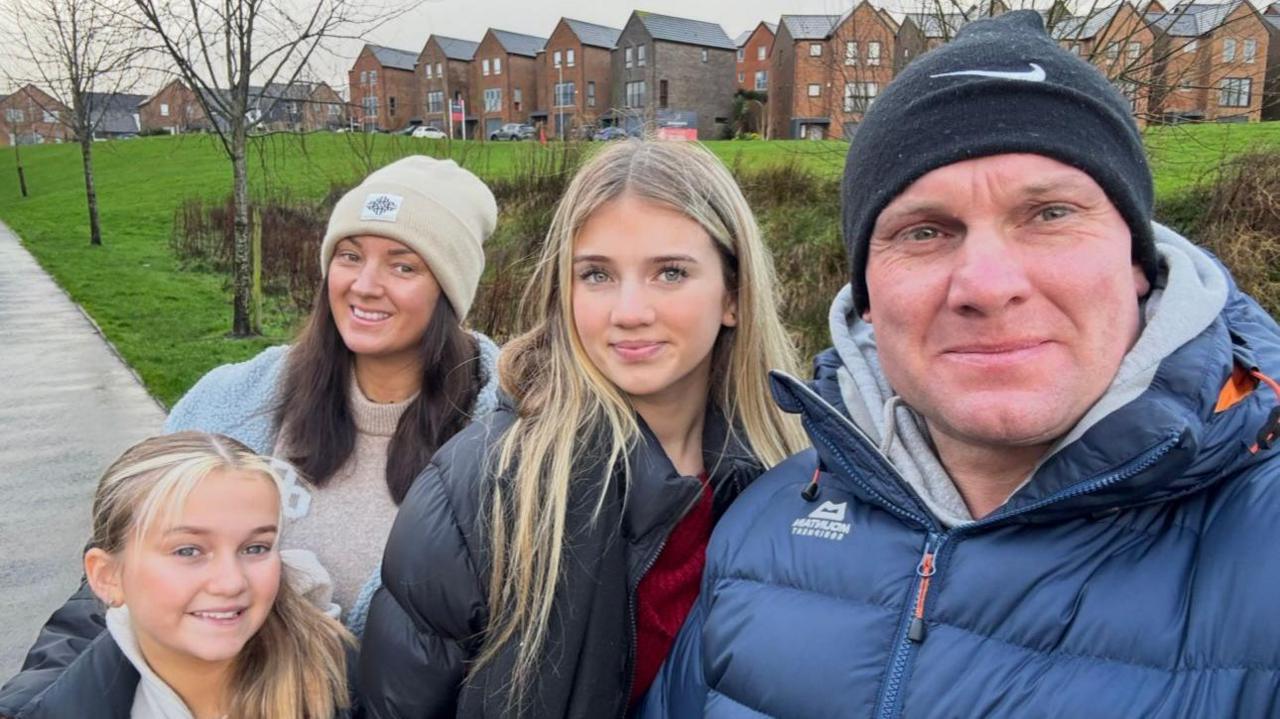 Betsy, who smiles and has a straw-coloured ponytail, poses with mum Rebecca, her older sister and dad in a group picture. They are near a green area in front of new housing development.