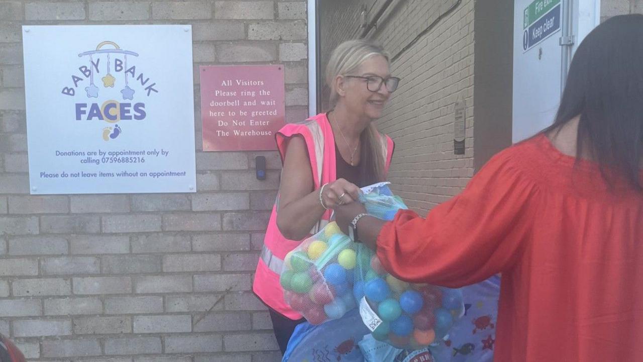 A bag of balls, being given to a woman, in a pink high-vis jacket, by an open door in a brick building. You can see the back of a woman in a red dress. 