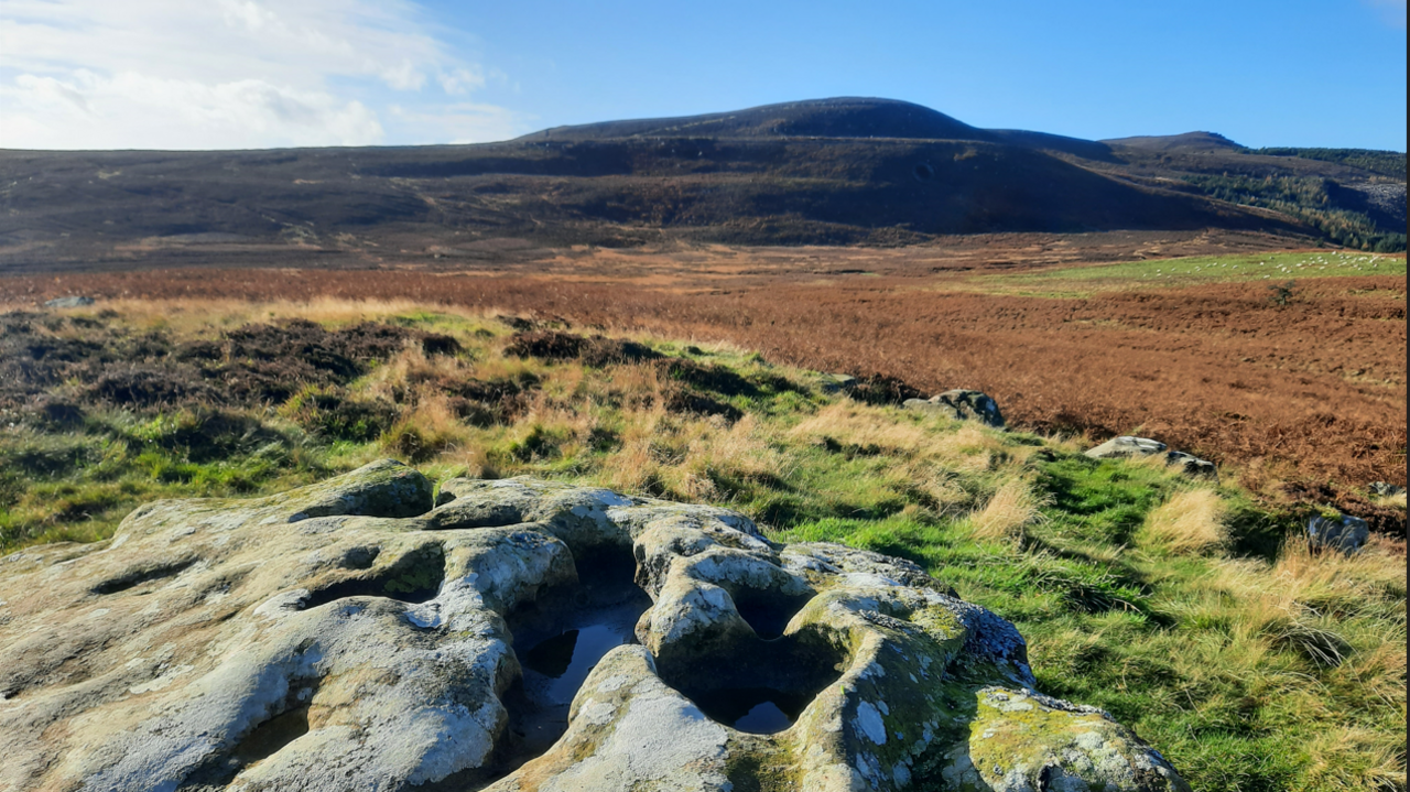 The Simonside Hills are in the centre rising in a dome shape and covered with brown bracken. In the foreground is a stone with water gathering in holes.