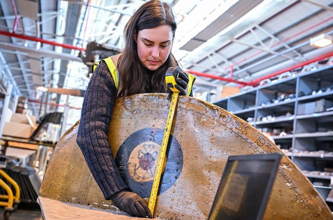 Someone measuring a large, mainly brown, object with a yellow tape measure. Shelves are in the background.
