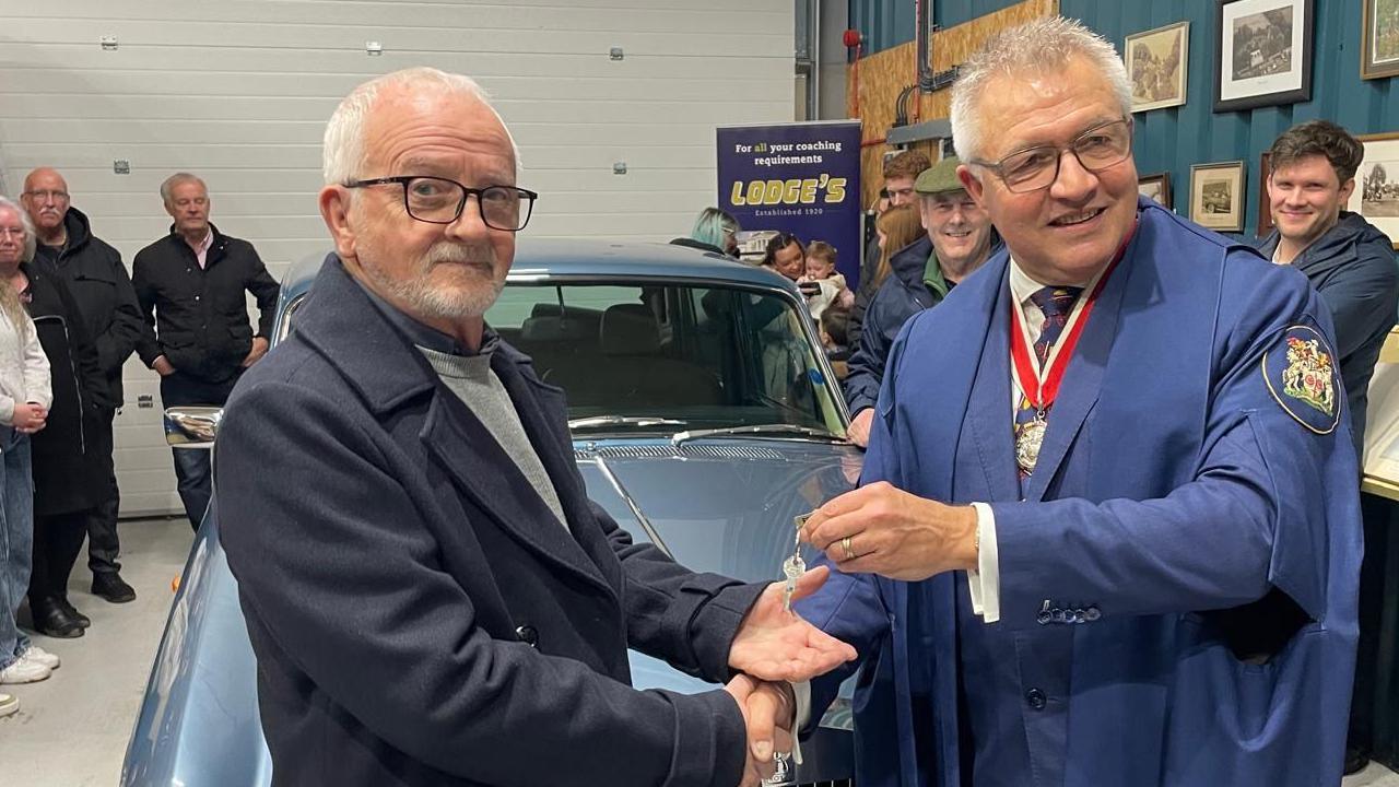 Mr Adams stands in front of his Rolls Royce. He shakes hands with Stephen Britt was he passes over the vehicle's keys in the other hand. The men smile as they pose for a photo. Mr Britt wears a blue uniform and has short grey hair with glasses.