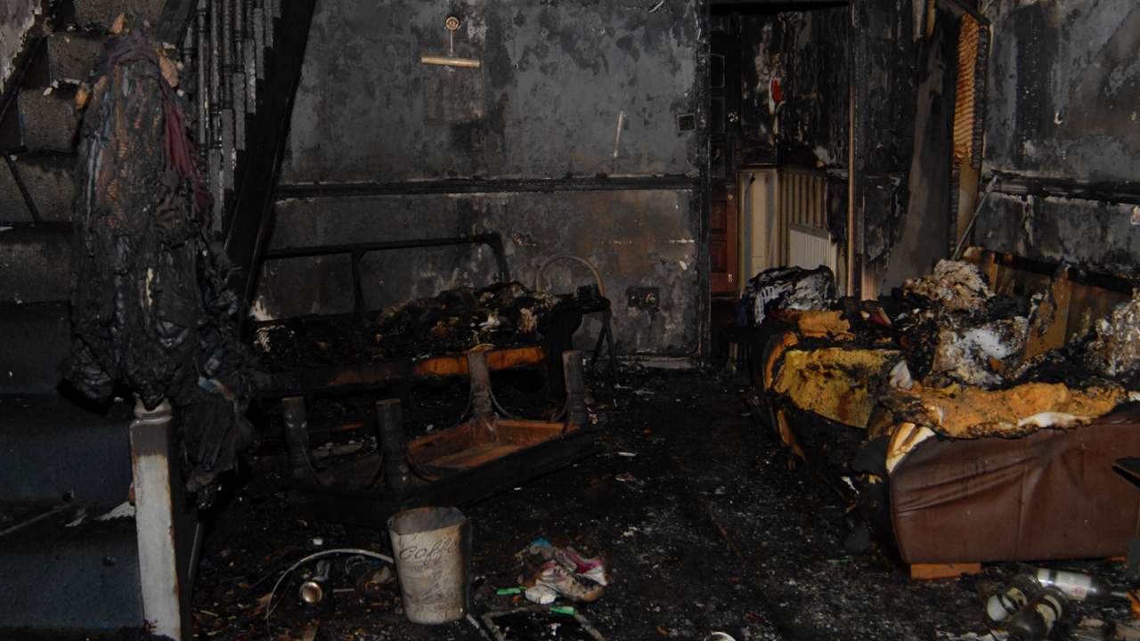 The burnt-out house shows the remains of a sofa to one side and a charred staircase at the other side of the room. The walls, floor and ceiling are all burnt and there is debris everywhere. Material is hanging off the walls. A destroyed table is turned upside down in the middle of the floor.