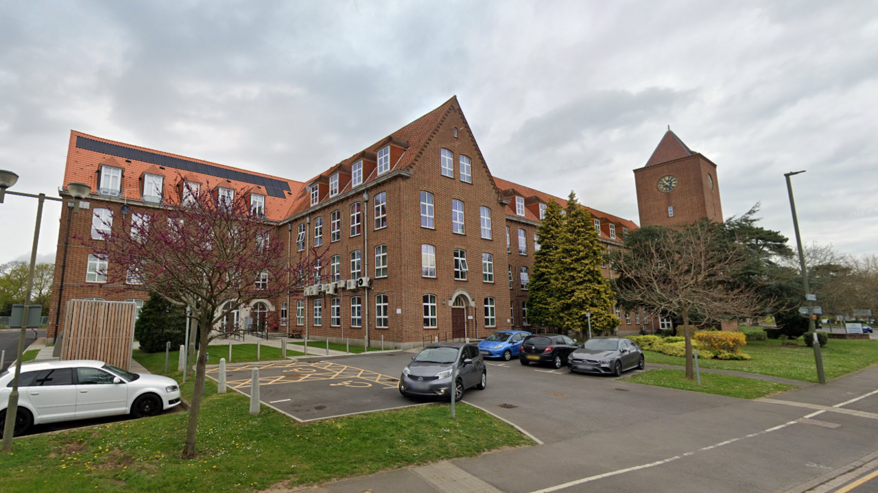 A large red brick building with several windows and a clock tower on the right hand side. The building has a car park in front of it with four black, grey and blue cars in it. Trees and grass are also in front of the building.