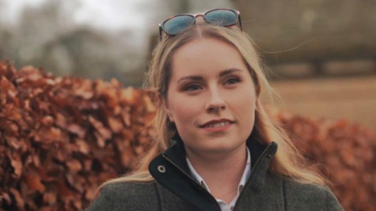 A young blonde woman looking off into the distance with a faint smile on her face wearing a grey tweet jacket. She has sunglasses on her head. In the background is a brown hedge, looking very autumnal.