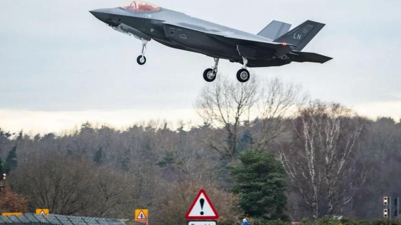 Dark gray US fighter jet with US Air Force livery. It is coming in to land at an airbase. In the background trees can be seen. There is a traffic sign below it.