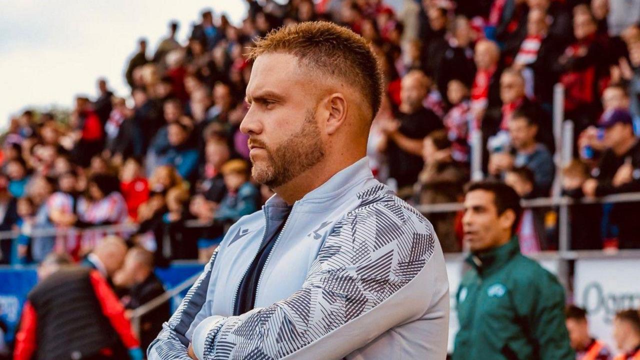 Nathan Rooney, wearing a grey jacket, stands with his arms folded in front of a crowded stand, with the picture taken from the left