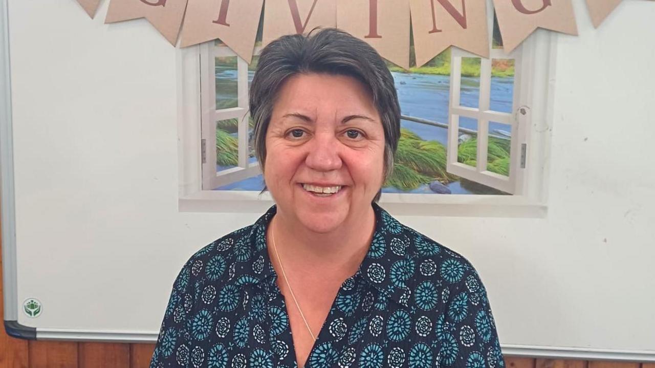 A smiling woman with dark grey hair and blue shirt stood in front of a wall at a Derbyshire care home