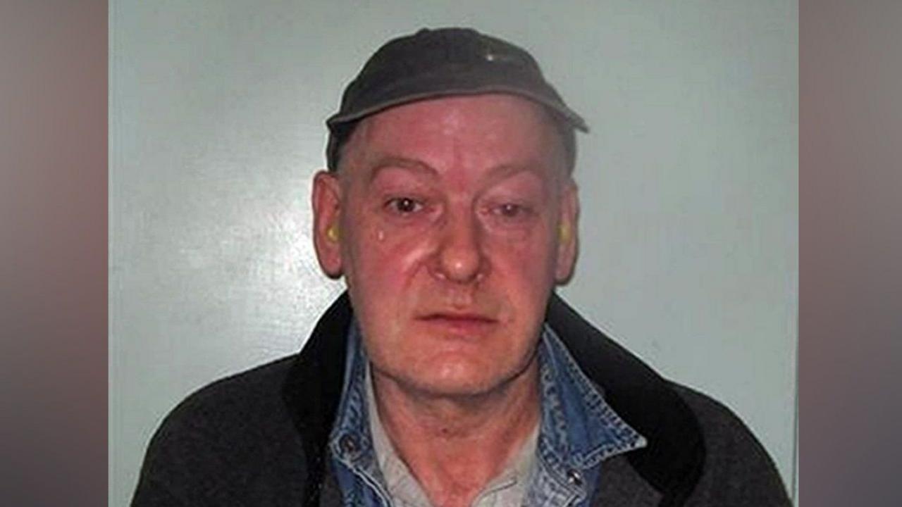 A custody-style photograph showing a Sweeney wearing a dark cap, denim shirt layered under a dark jacket and a light-coloured top, standing against a plain light background.