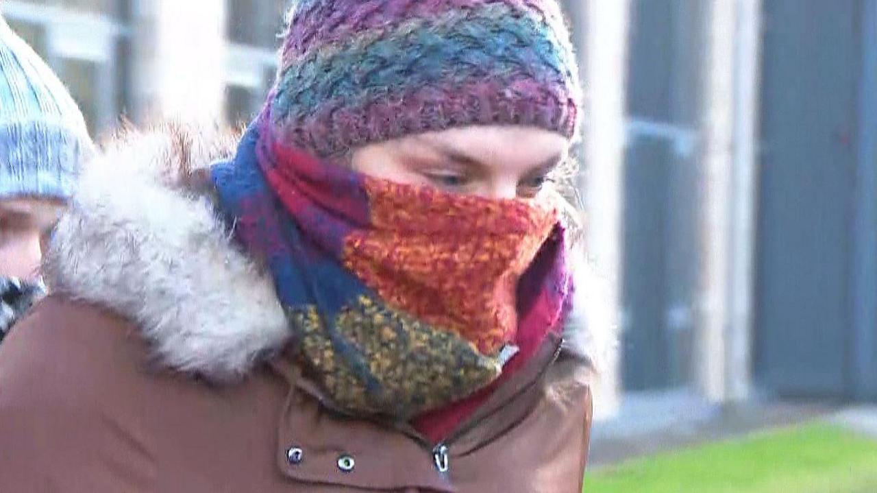 Dr Beth Upton is shown wearing a brown fur-lined jacket while leaving the tribunal office in Dundee. The doctor's face is hidden by a multi-coloured scarf and a purple and blue bobble hat. 