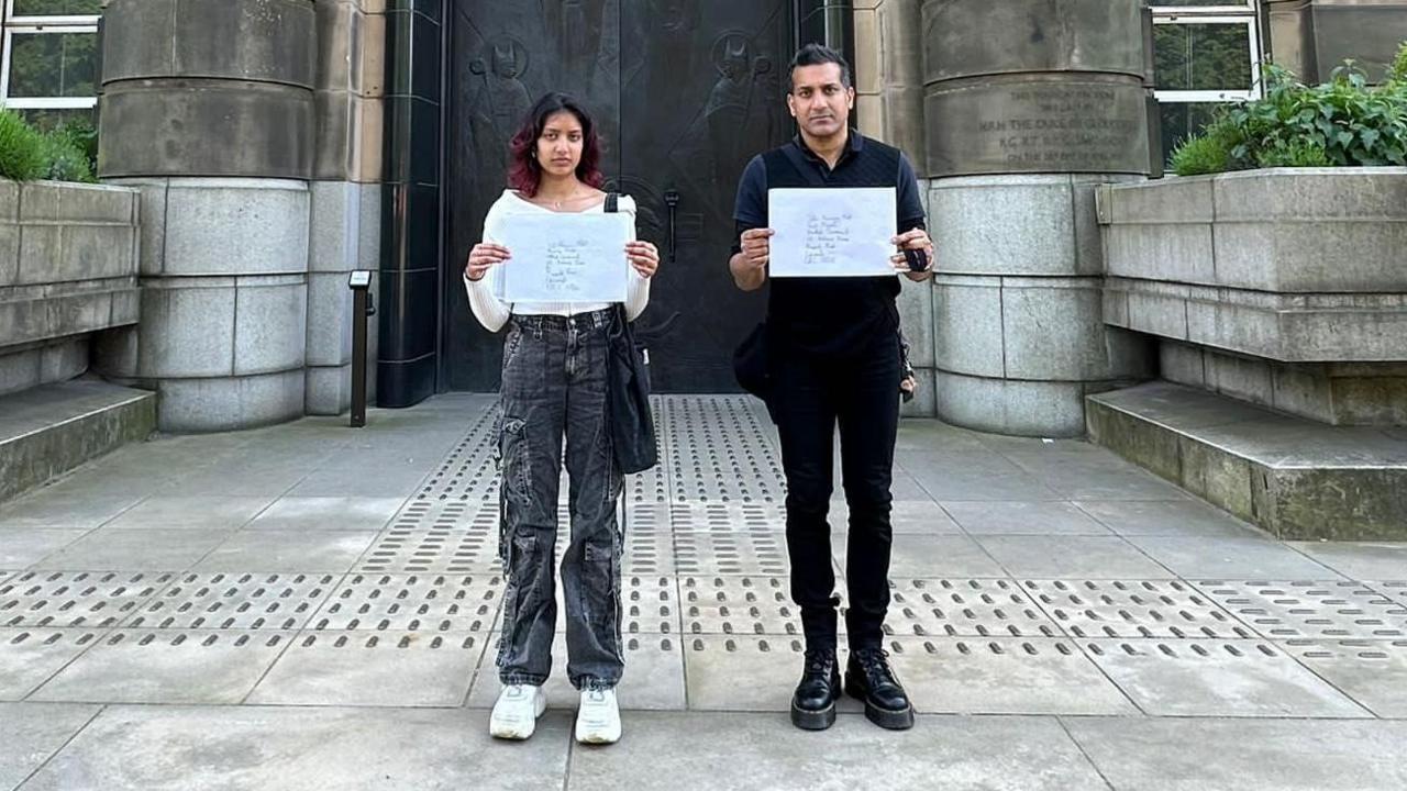 A man, Wilson Chowdhry, and a woman, Hannah Chowdhry, stand in front of an ornate stone building. They are each holding a piece of paper in their hands and looking seriously into the camera.